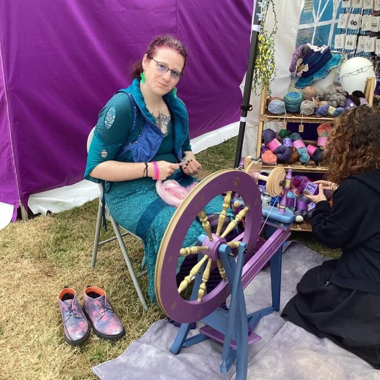 adhcrafts nessi A woman spinning yarn at a craft fair, with a colourful backdrop and another person working nearby.